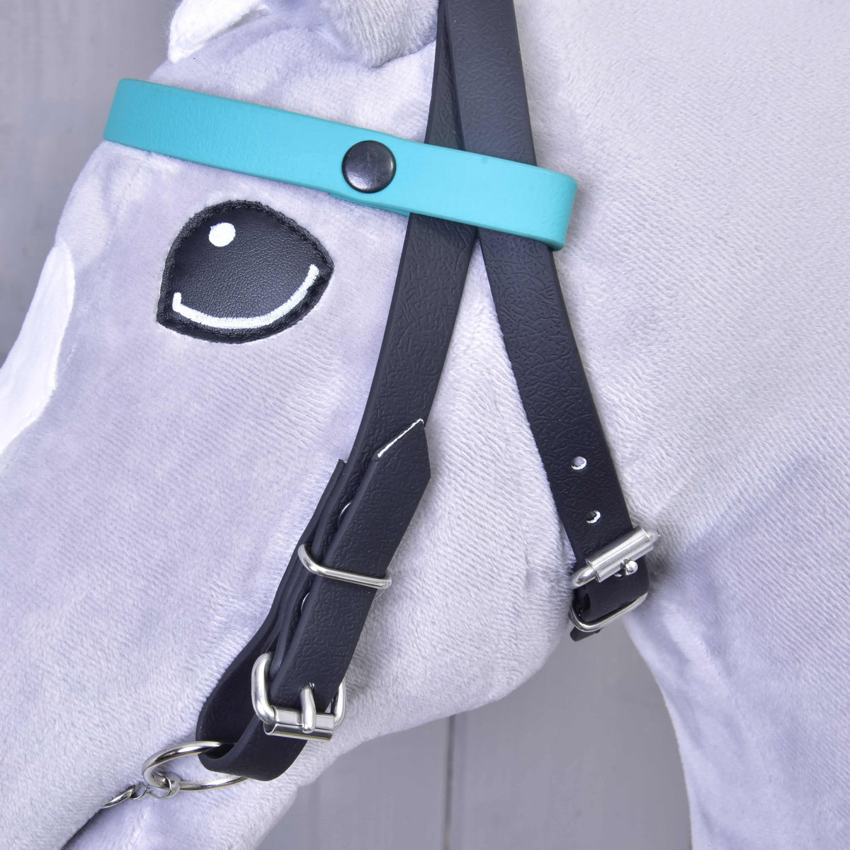 Bližnji posnetek enobarvnega turkiznega čelnika s črnim pritiskačem na črni uzdi sivega Hobby Horse konja