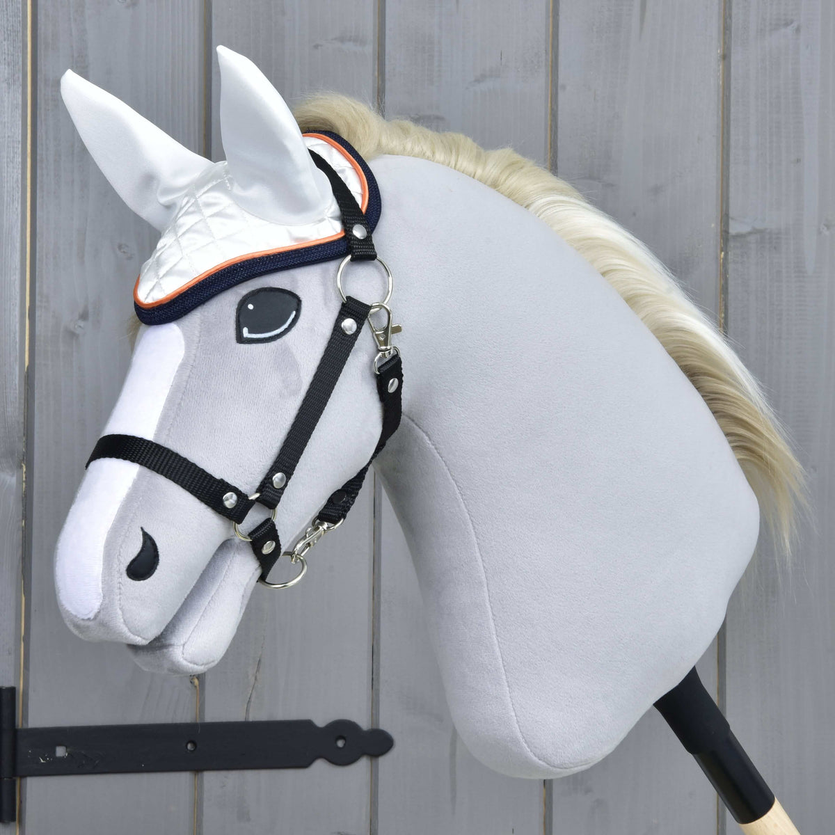 Siv plišast Hobby Horse konj s smetanasto grivo in črno oglavko nosi bele naušnike z oranžno in temno modro obrobo