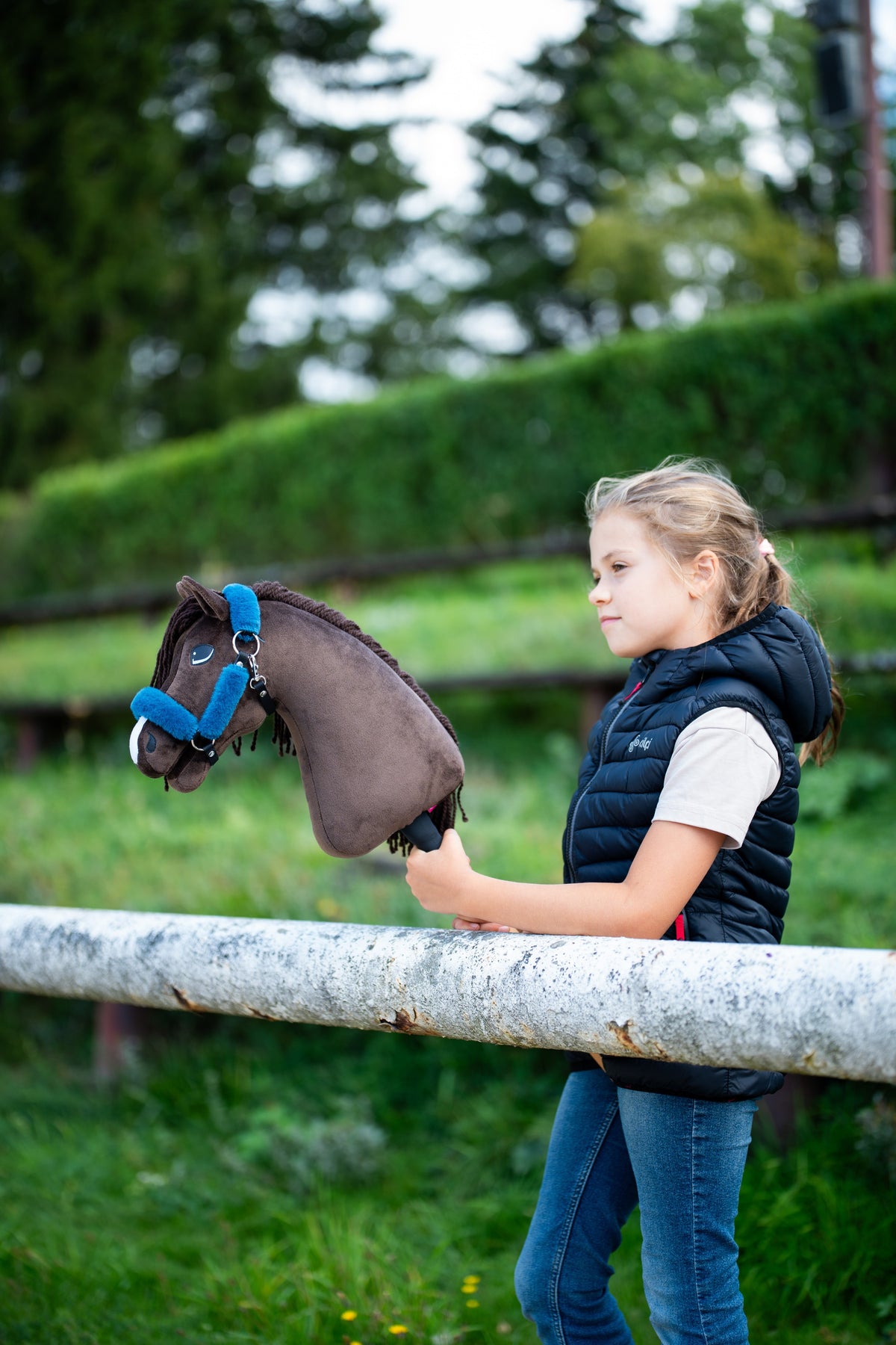 Deklica v temno modrem podloženem brezrokavniku ob beli ograji drži temno rjavega Hobby Horse konja z modro oglavko s krznom, za njo živa meja