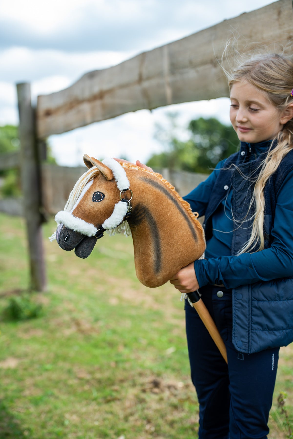 Girl in a navy gilet by a paddock fence holds a light brown Hobby Horse with a dark stripe, wearing the white fur halter