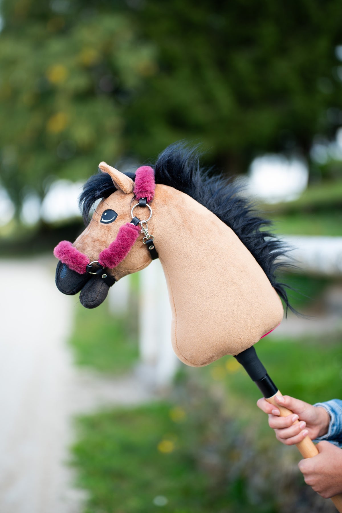 Hobby Horse konj Cookie v črni oglavki z malinovo roza krznom in kosmato črno grivo, dvignjen pred drevesi