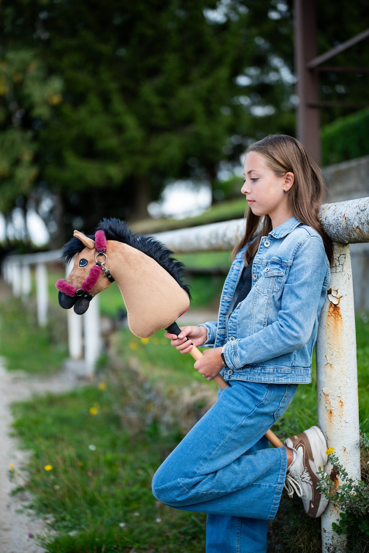 Deklica v jakni iz džinsa, naslonjena na belo ograjo, drži svetlo rjavega Hobby Horse konja z malinasto oglavko s krznom