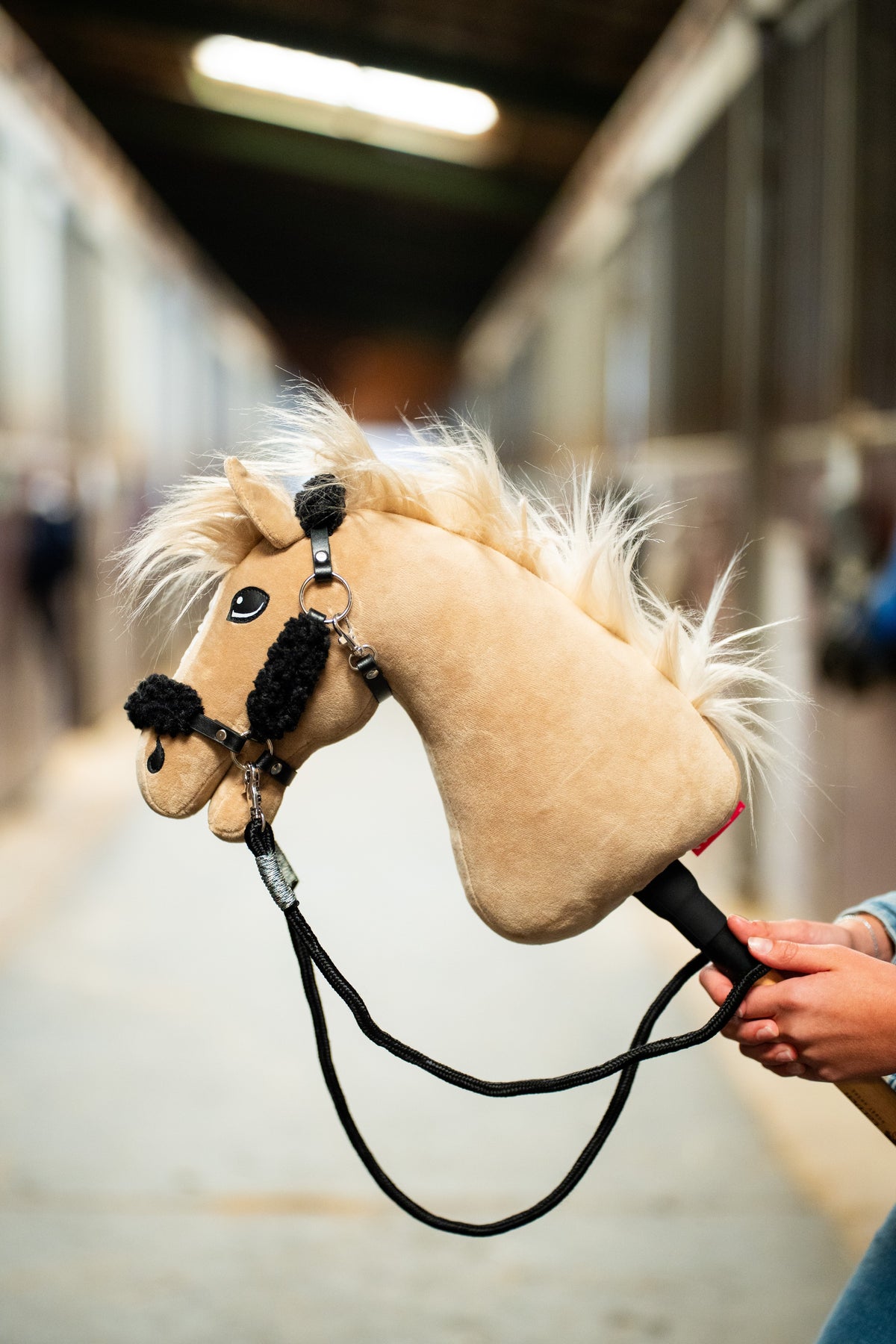 Hobby Horse Dancing Lady with a fluffy cream mane in a black fur halter, the black braided reins clipped on, stable aisle