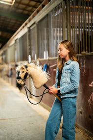 Deklica v jeans jakni in kavbojkah v hlevskem hodniku drži smetanastega Hobby Horse konja s črno oglavko s krznom za črne vajeti