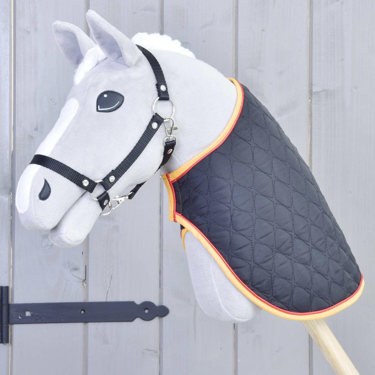 Grey Hobby Horse with a black halter wearing the black quilted blanket with gold and red trim on its neck
