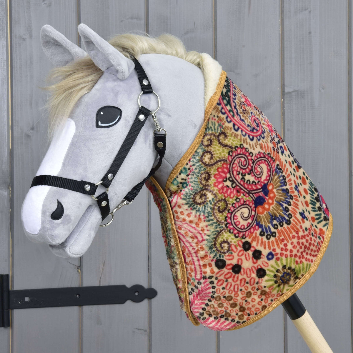 Siv plišast Hobby Horse konj s črno oglavko nosi pisano vzorčasto pregrinjalo z roza, zelenimi in temno modrimi vrtinci