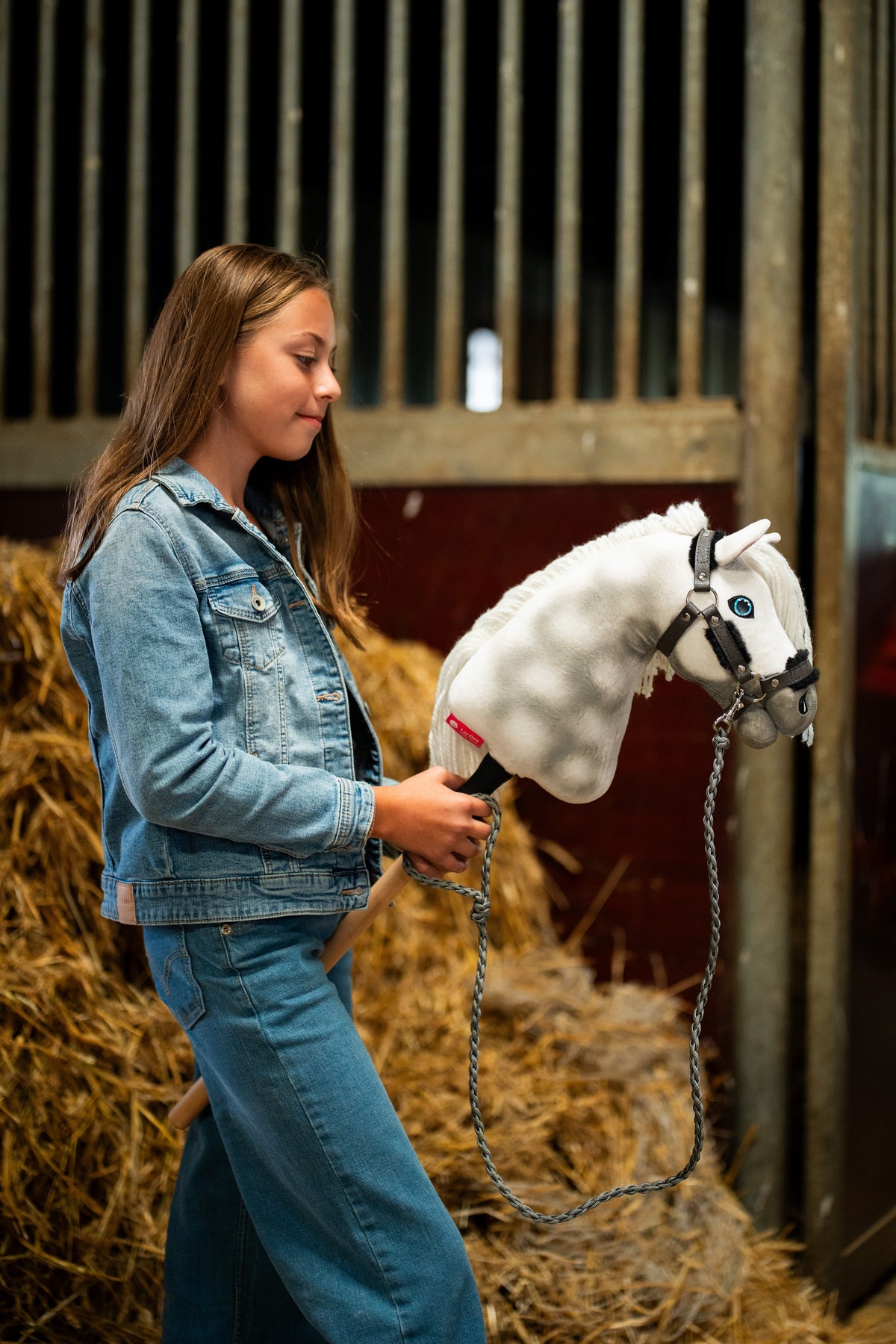 Deklica v jeans jakni ob balah slame drži Hobby Horse konja Elektra, tukaj s temno sivo oglavko s krznom in pletenim sivim povodcem