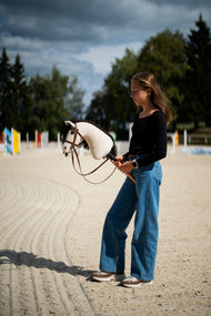 Deklica na peščenem jahališču z barvitimi ovirami drži kremnega Hobby Horse konja z rjavo kompletno uzdo z nosnikom