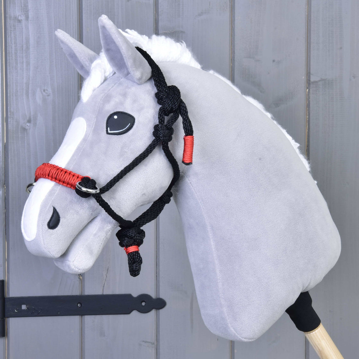 Siv plišast Hobby Horse konj z belo grivo nosi črno-rdečo vrvično oglavko, pred sivo leseno steno