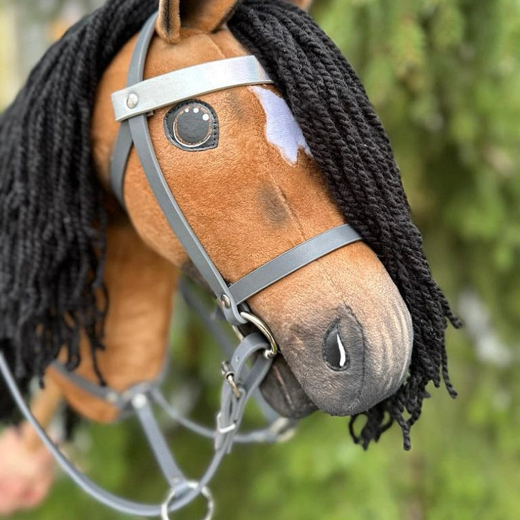 Bližnji posnetek ročno izdelanega Hobby Horse konja s črno grivo iz preje, rjavim telesom, belo liso nad enim očesom in sivo uzdo pred zamegljenim zelenim ozadjem v naravi, popoln dodatek za dogodivščine v športu Hobby Horsing.