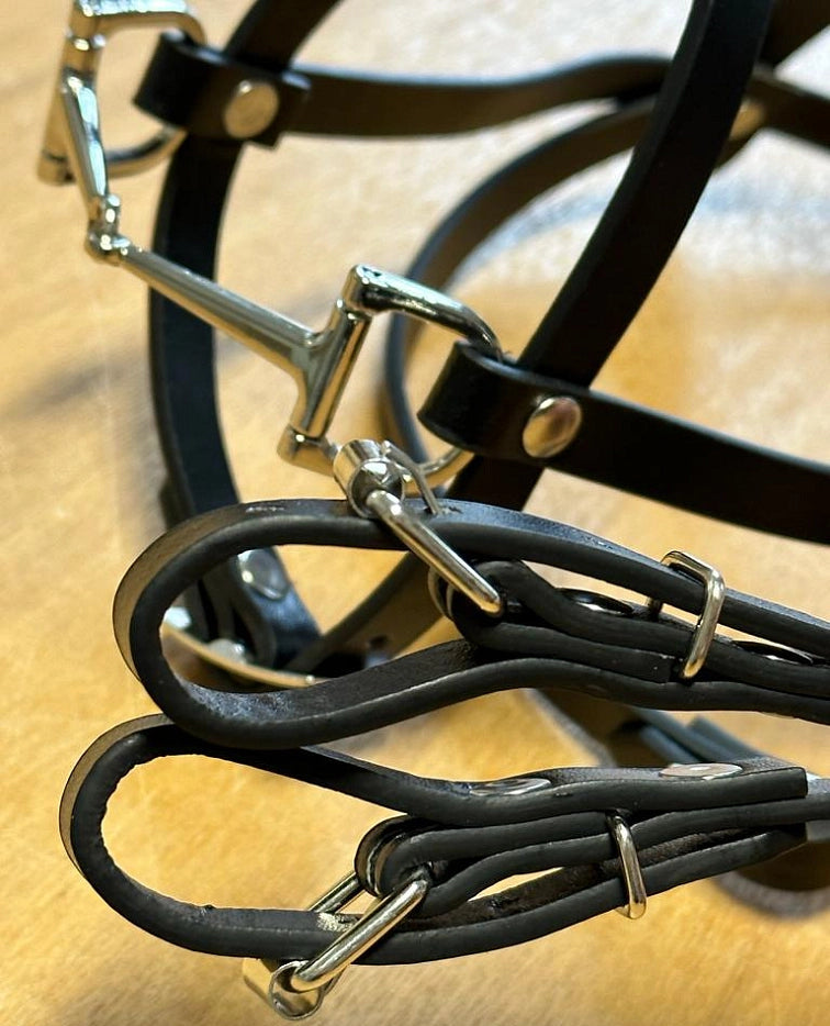 Bližnji posnetek črne uzde in vajeti iz umetnega usnja s svetlečimi srebrnimi kovinskimi zaponkami in obročki, kot nalašč za opremo za Hobby Horsing, na svetlo rjavi podlagi.
