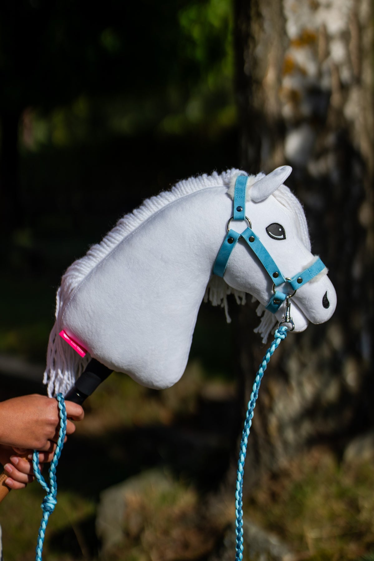 Hobby Horse konj Lady z belo resasto grivo v svetlo modri oglavki, pri bradi pripet svetlo moder pleten povodec, sončen vrt