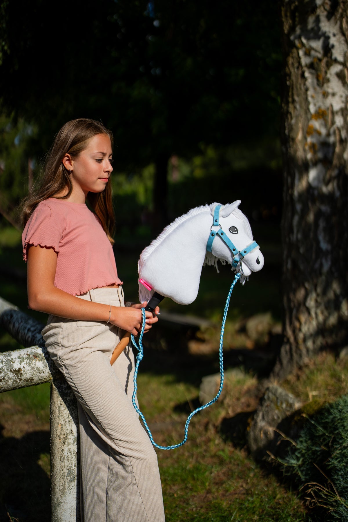 Deklica v roza majici ob brezi drži belega Hobby Horse konja s svetlo modro oglavko in povodcem v zanki