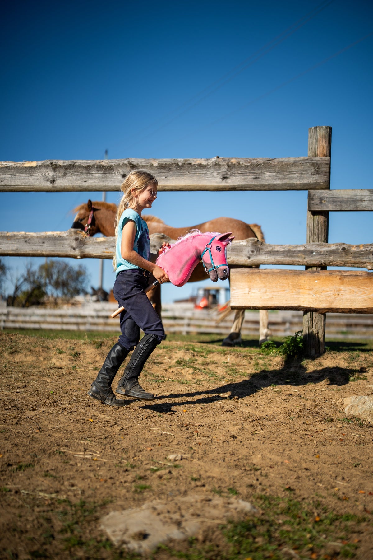 Nasmejana deklica v jahalnih škornjih vodi Hobby Horse konja Pinkie, tu s turkizno oglavko, čez izpust mimo lisjaka