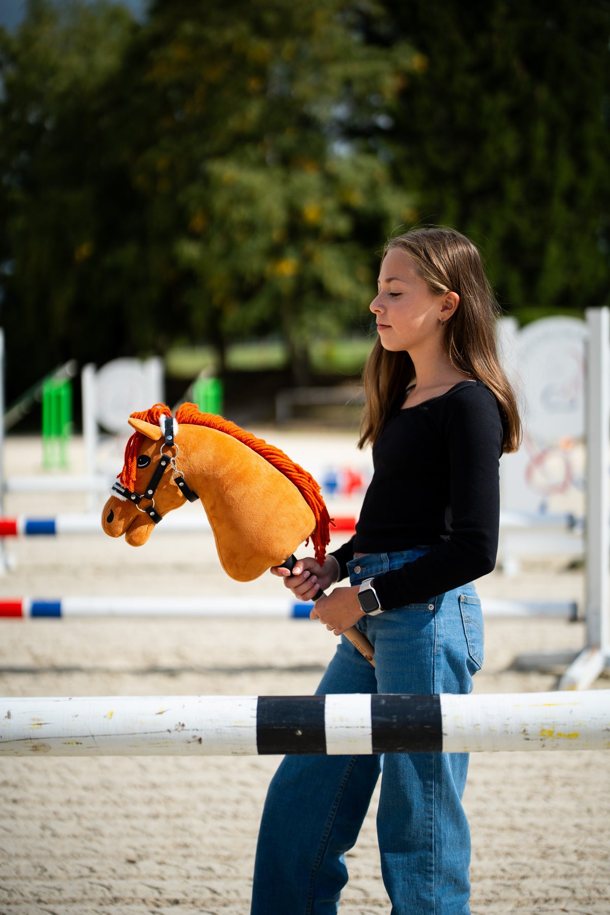 Deklica v črni majici na peščenem jahališču z ovirami drži Hobby Horse konja lisjaka s črno oglavko z belim krznom