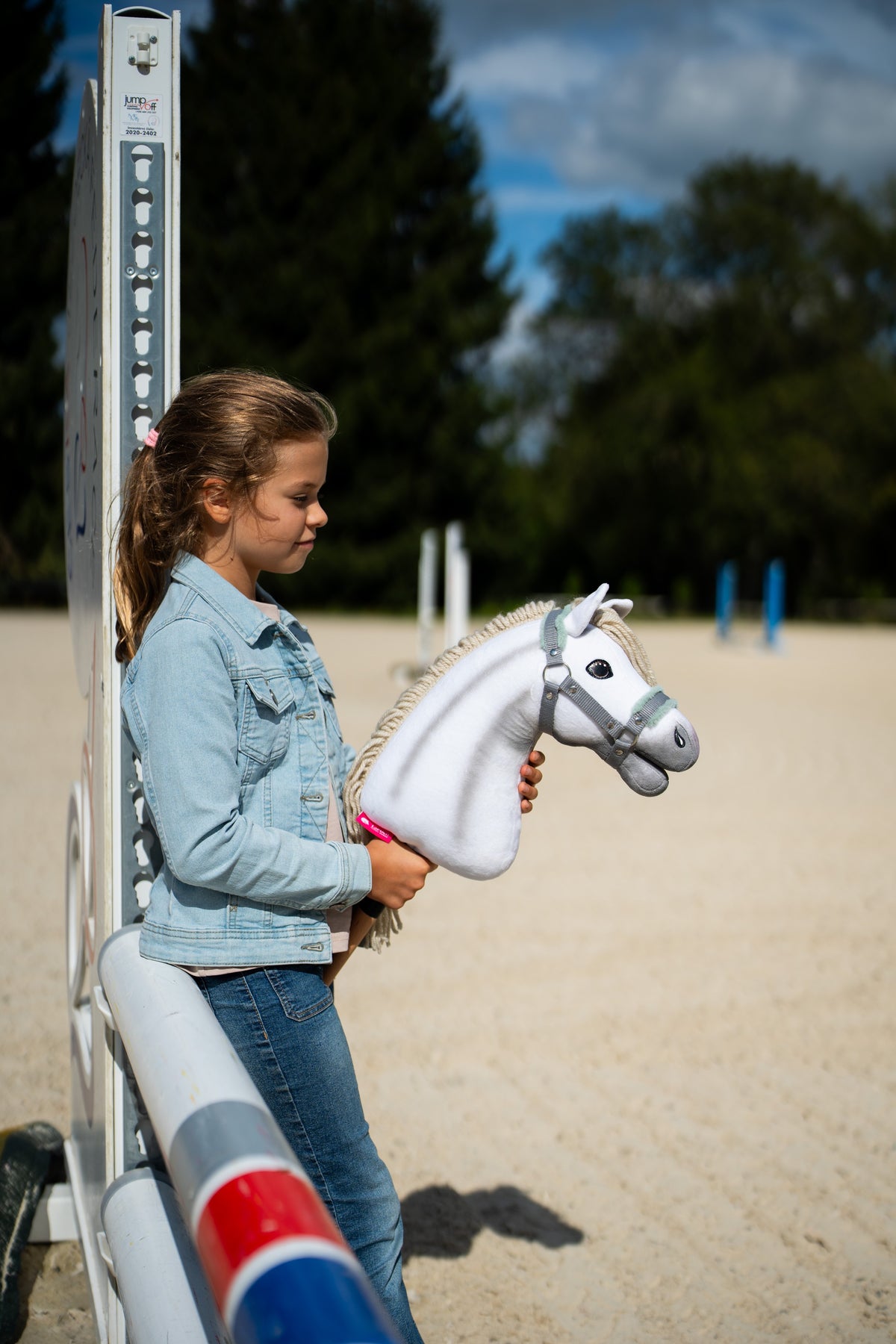 A girl in a denim jacket leans on a jump stand in a sand arena, holding a white Hobby Horse in the grey halter with mint fur
