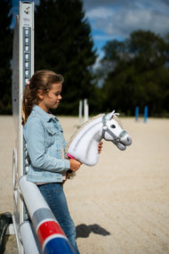 Deklica v jeans jakni se na peščenem jahališču naslanja na stojalo ovire in drži belega Hobby Horse konja v sivi oglavki z metino zelenim krznom