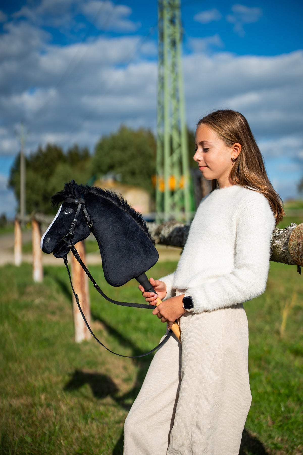 Girl in a white fluffy jumper in a sunny meadow holding Hobby Horse Loki in a black bridle with reins