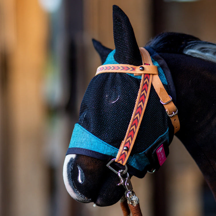 Bližnji posnetek konja v modri mrežasti maski proti mrčesu z zaščito za ušesa in rjavi usnjeni oglavki z rdečim vzorcem puščic, ki spominja na opremo za Hobby Horsing. Ozadje je mehko zamegljeno.