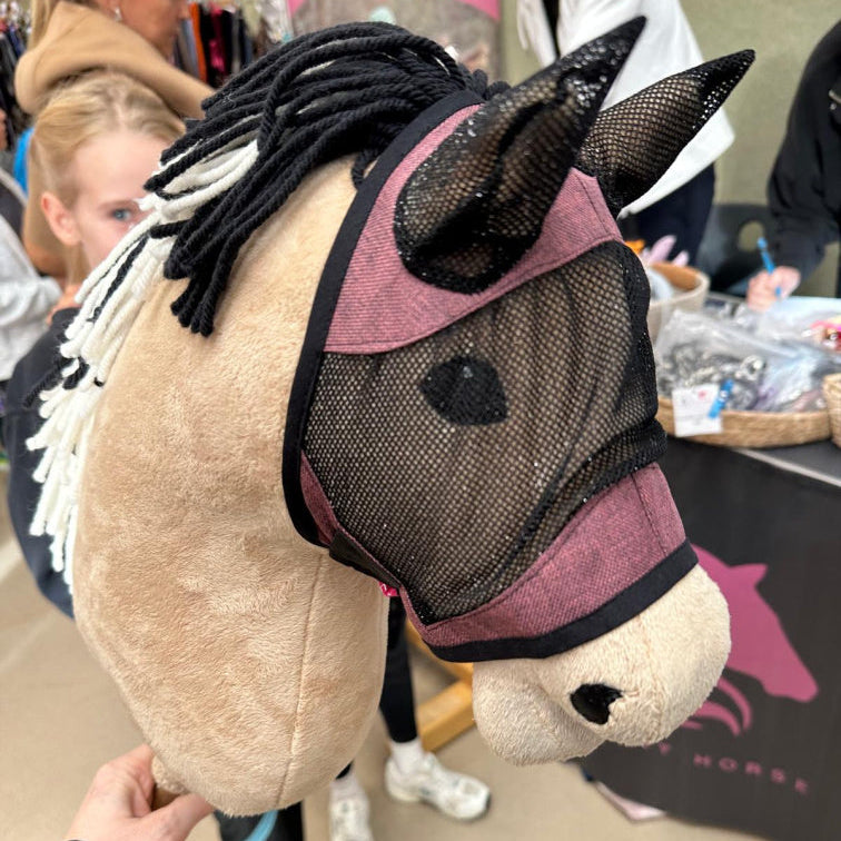 Bližnji posnetek ročno izdelanega Hobby Horse konja z mrežasto masko in črno-belo grivo iz preje. V zamegljenem ozadju ljudje in stojnica z izdelki s konjskimi motivi prikazujejo ustvarjalni duh športa Hobby Horsing.