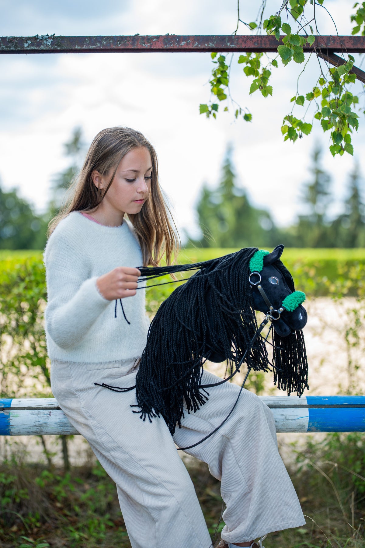 Girl in a white jumper sitting on a jump pole, gathering the reins of the black Hobby Horse in a green fur halter