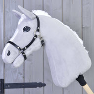 Siv plišast Hobby Horse konj s puhasto belo grivo in liso nosi črno oglavko iz umetnega usnja, pred sivo leseno steno