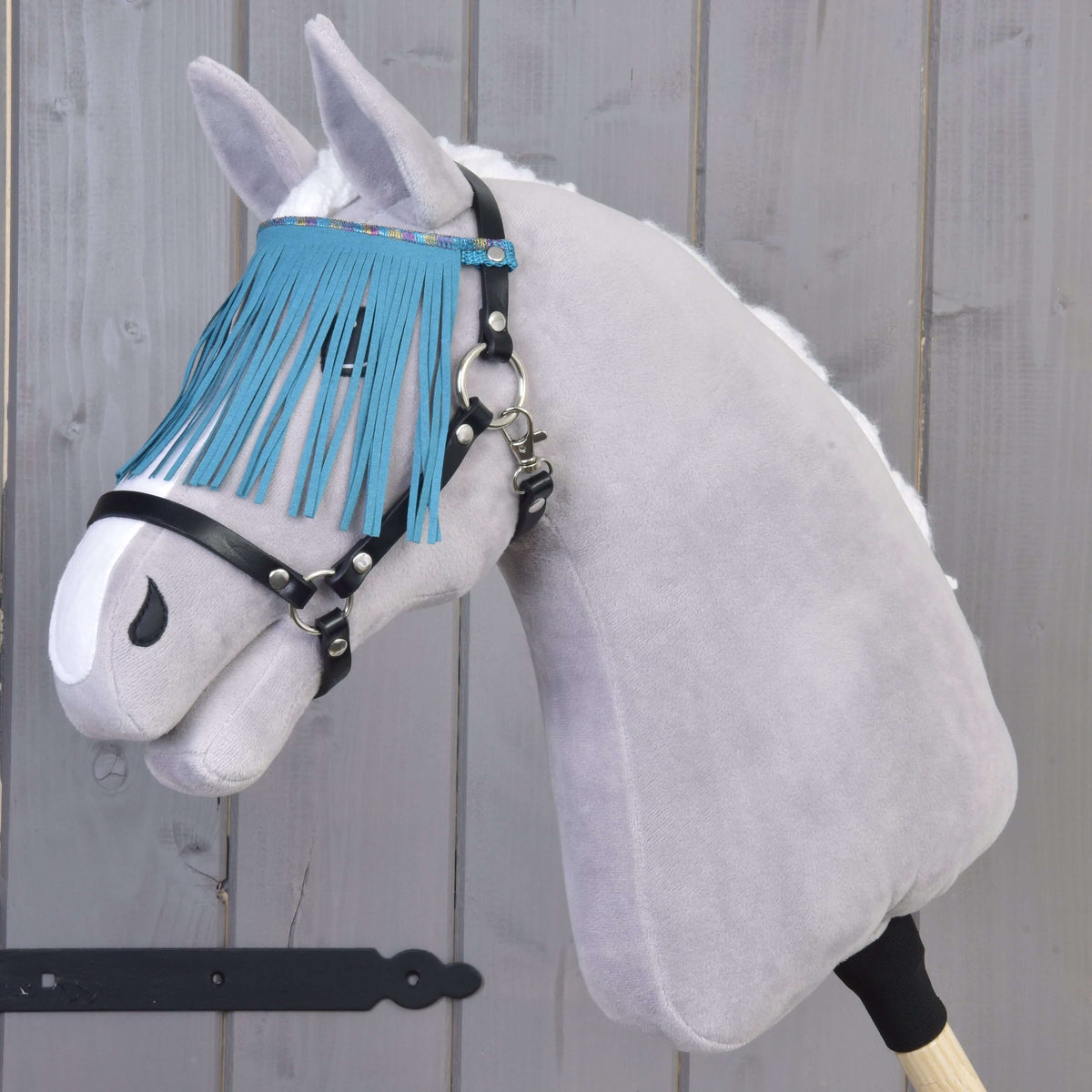 Grey plush Hobby Horse with a white mane wearing the black halter, turquoise fringes hanging over its eyes