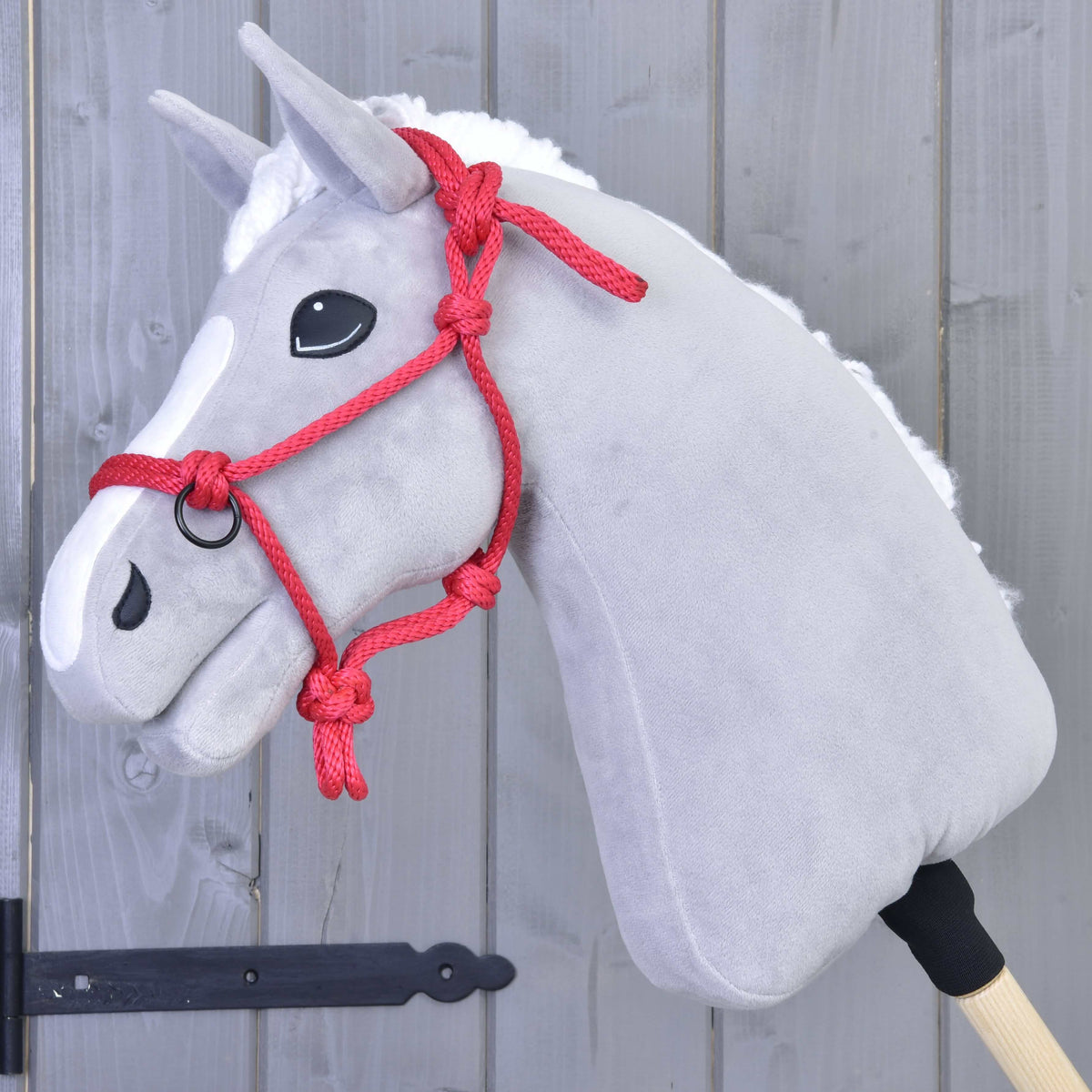 Grey plush Hobby Horse with white blaze and mane wearing the red rope halter against a grey wooden wall