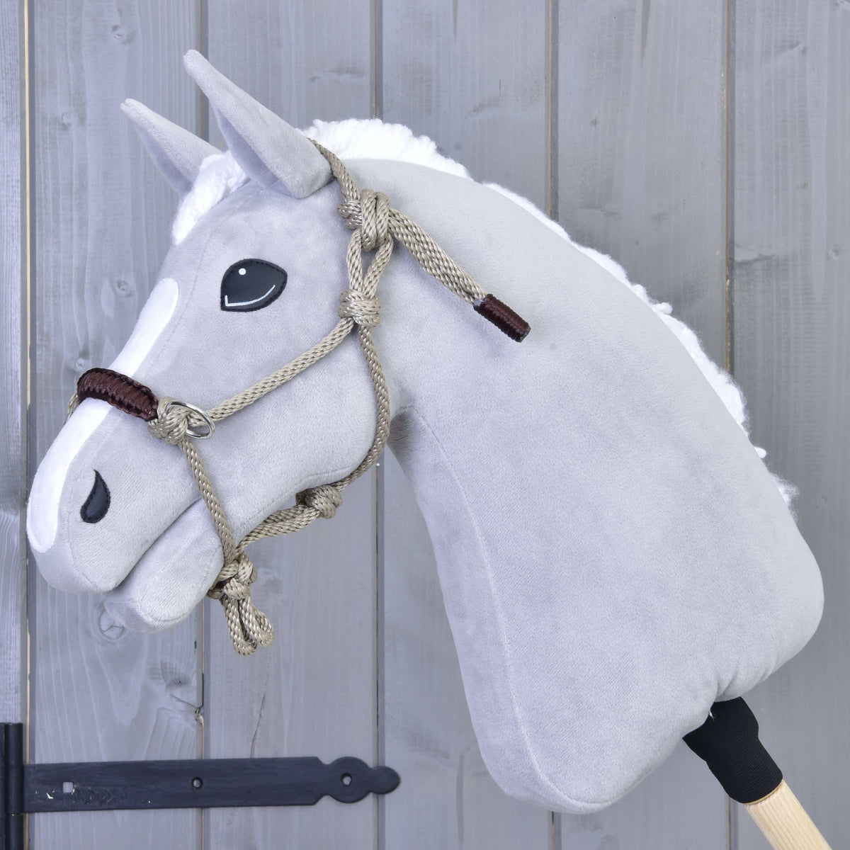 Grey plush Hobby Horse with a white mane wearing the beige rope halter with brown noseband in front of a grey wooden wall