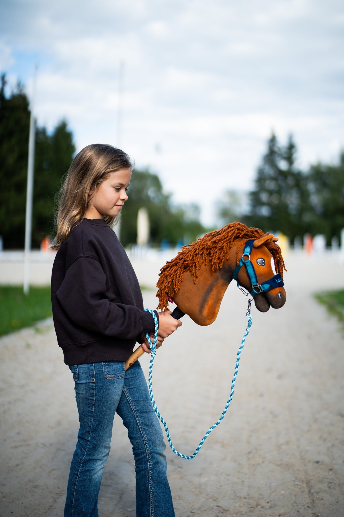 Young girl in a dark sweatshirt walking Hobby Horse Pippa in a teal halter and blue lead rope on a sandy track