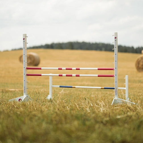 Belo-rdeče progasti drogovi za skakanje, postavljeni na travniku, kot nalašč za Hobby Horsing, v ozadju bale sena in drevesa pod oblačnim nebom.
