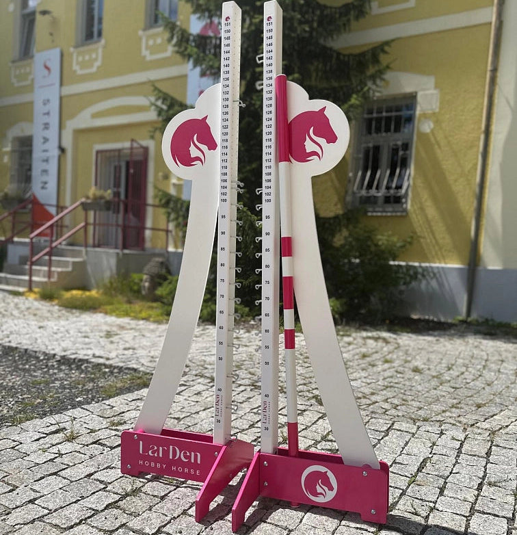 Dve belo-roza nastavljivi stojali ovir za Hobby Horsing, postavljeni na tlakovani poti na prostem, v ozadju rumena stavba s stopnicami in napisom.