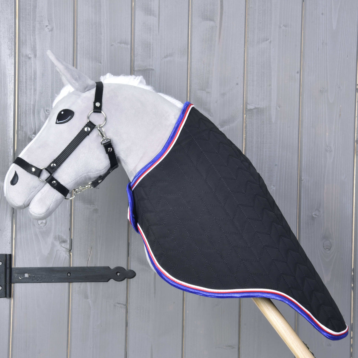 Side view of a grey Hobby Horse in the black quilted travel rug, which covers the whole neck down to the wooden stick