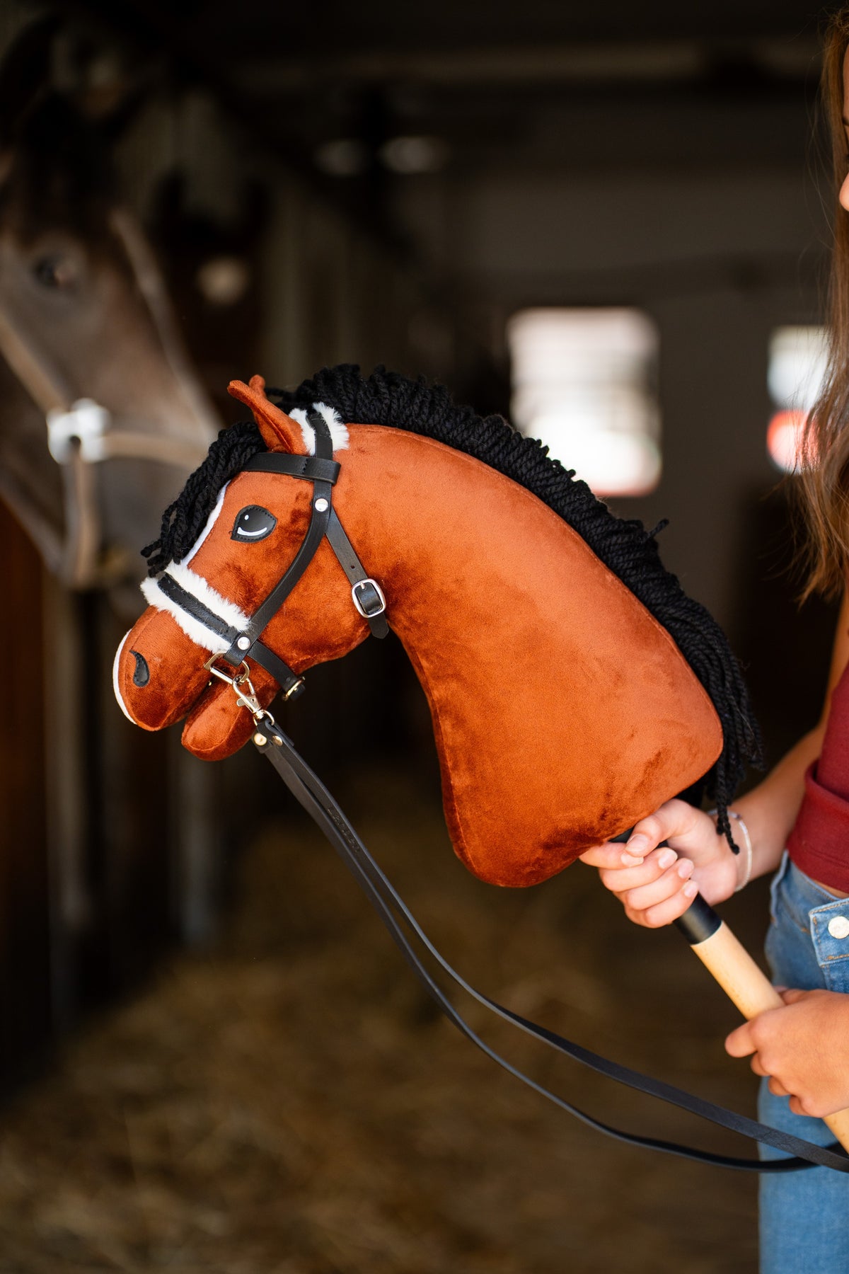 Hobby Horse konj Rocky kostanjeve barve s kodrasto črno grivo iz preje v črni uzdi z belim krznom in dolgimi vajetmi, zadaj rjavec