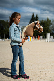 Girl in a denim jacket on a sand arena with jumps, holding a light brown Hobby Horse in the cream fur halter