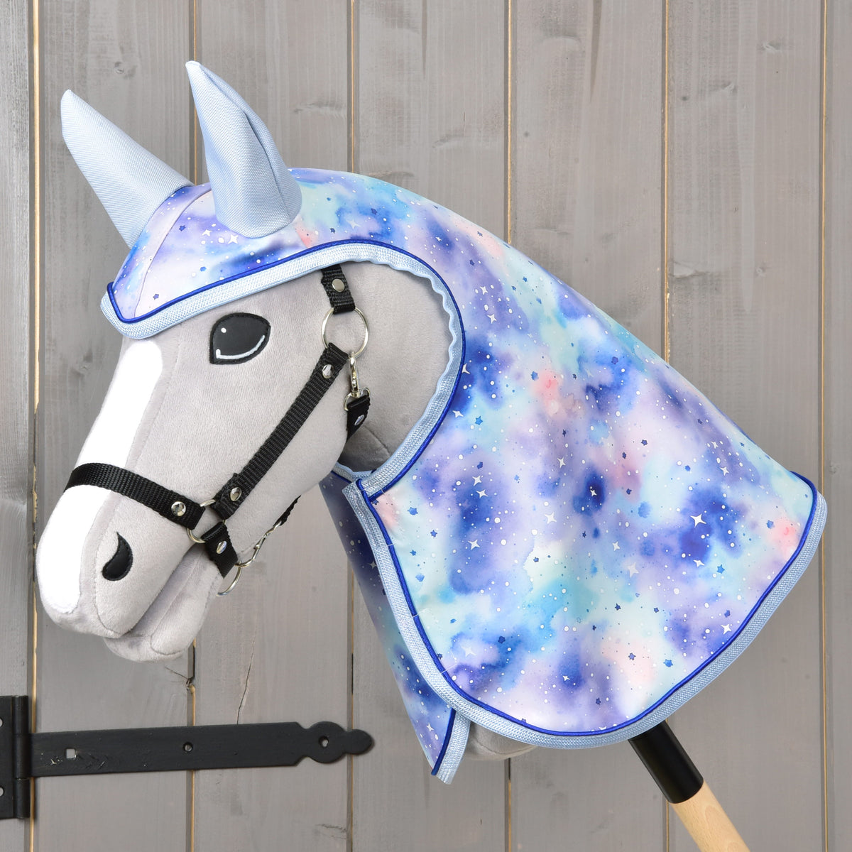 Siv Hobby Horse konj s črno oglavko nosi dežno pregrinjalo Space s kapuco in svetlo modrimi ušesi v modro-vijoličnem vzorcu galaksije