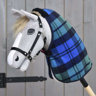 Grey plush Hobby Horse in a black halter wearing the blue and green tartan stable blanket against a grey wooden wall