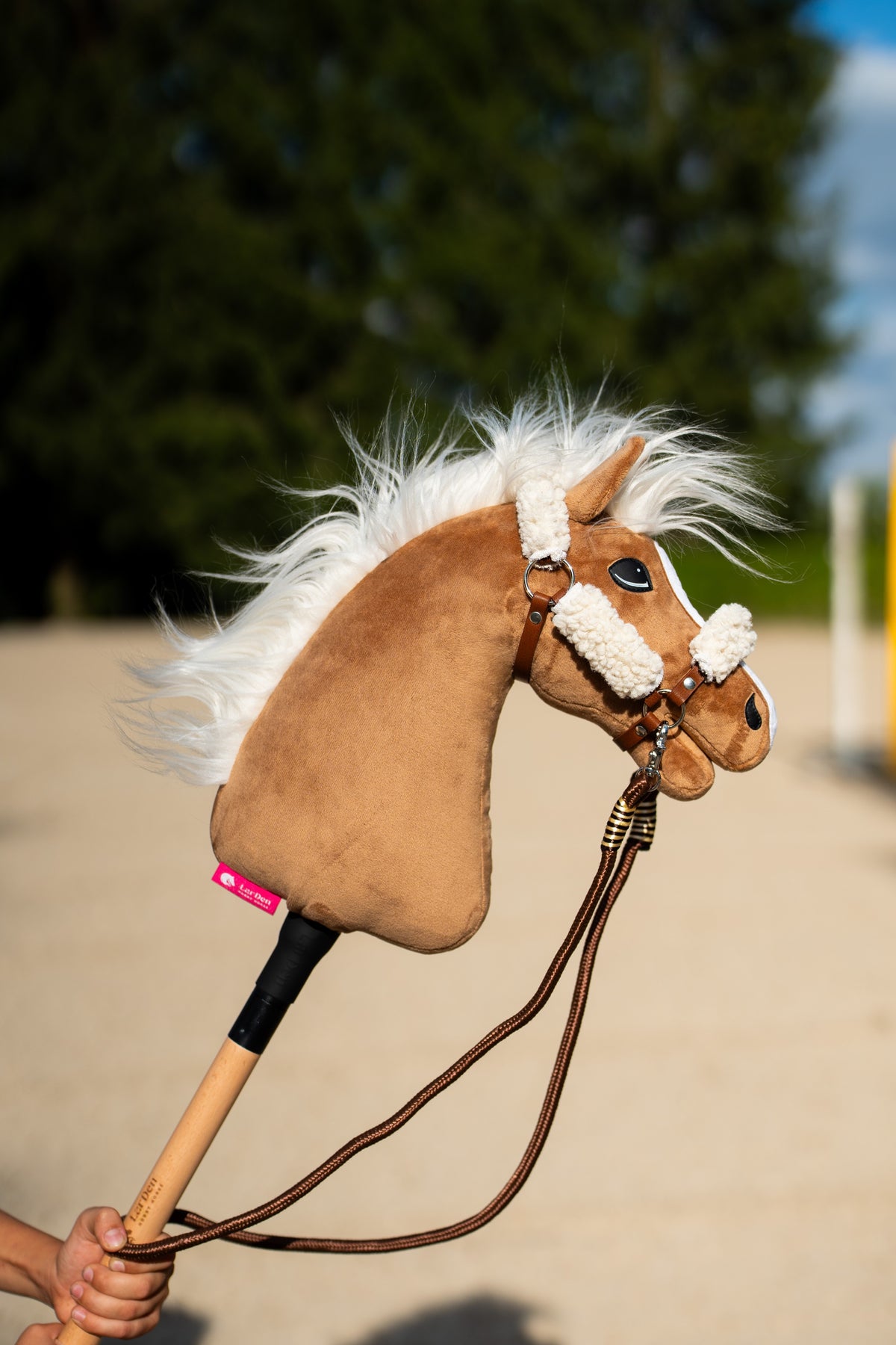 Hobby Horse konj Star v rjavi oglavki s kremnim krznom, rjave vrvične vajeti drži otroška roka na sončnem jahališču