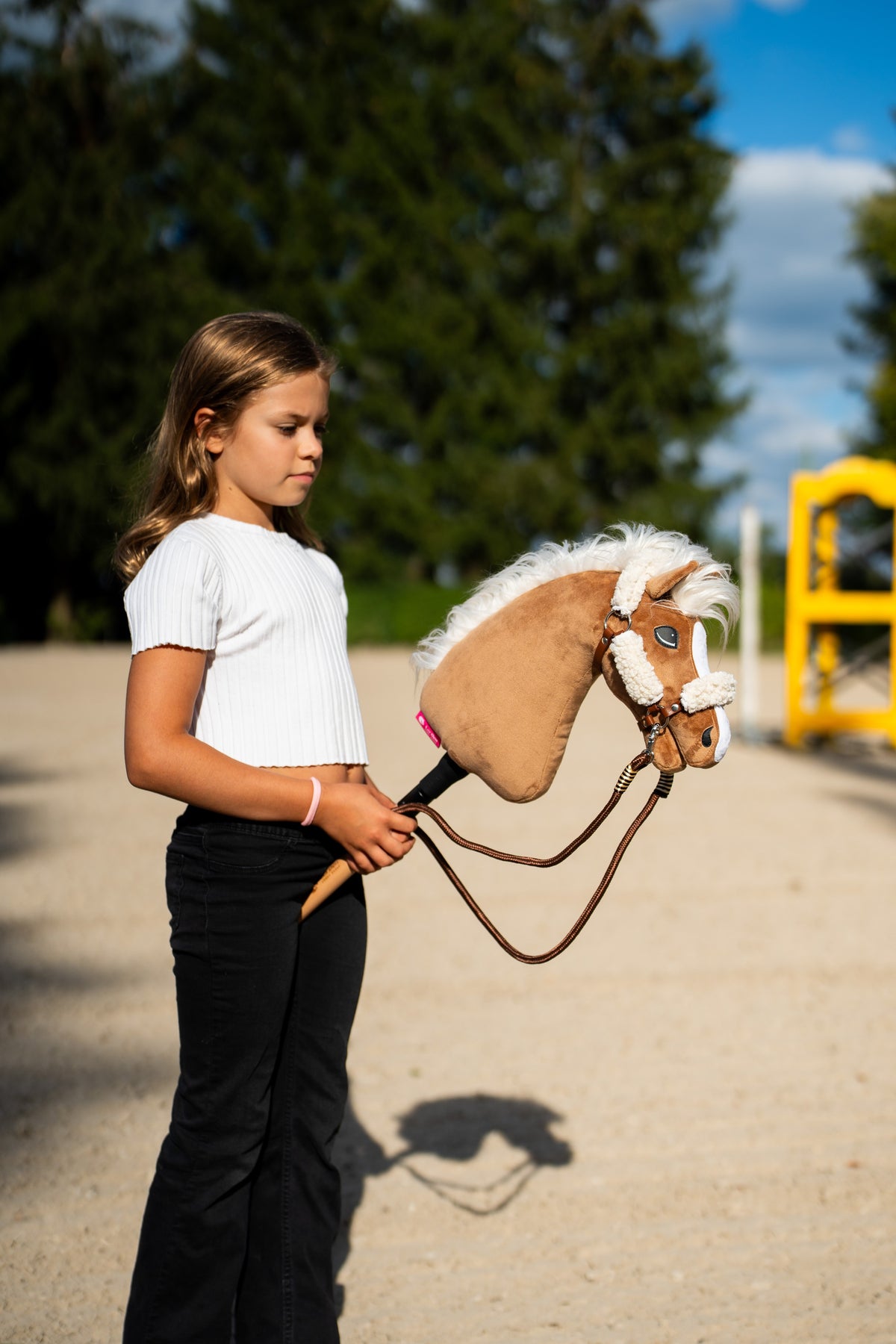 Deklica v beli majici na peščenem jahališču drži Hobby Horse konja Star, tukaj z rjavo oglavko s smetanastim krznom in rjavimi vrvičnimi vajetmi
