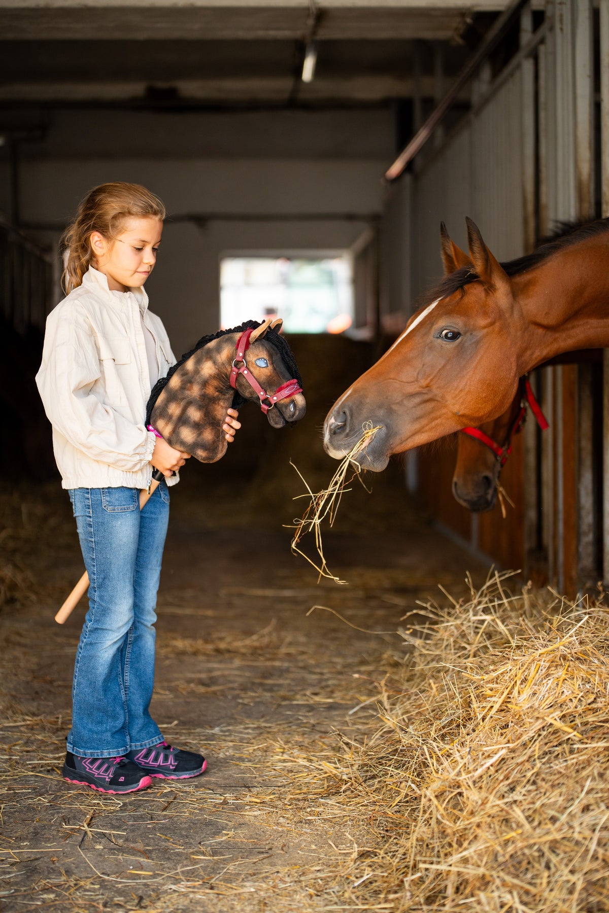Deklica v hlevu drži rjavo jabolkastega Hobby Horse konja z bordo oglavko s krznom ob rjavcu, ki je seno