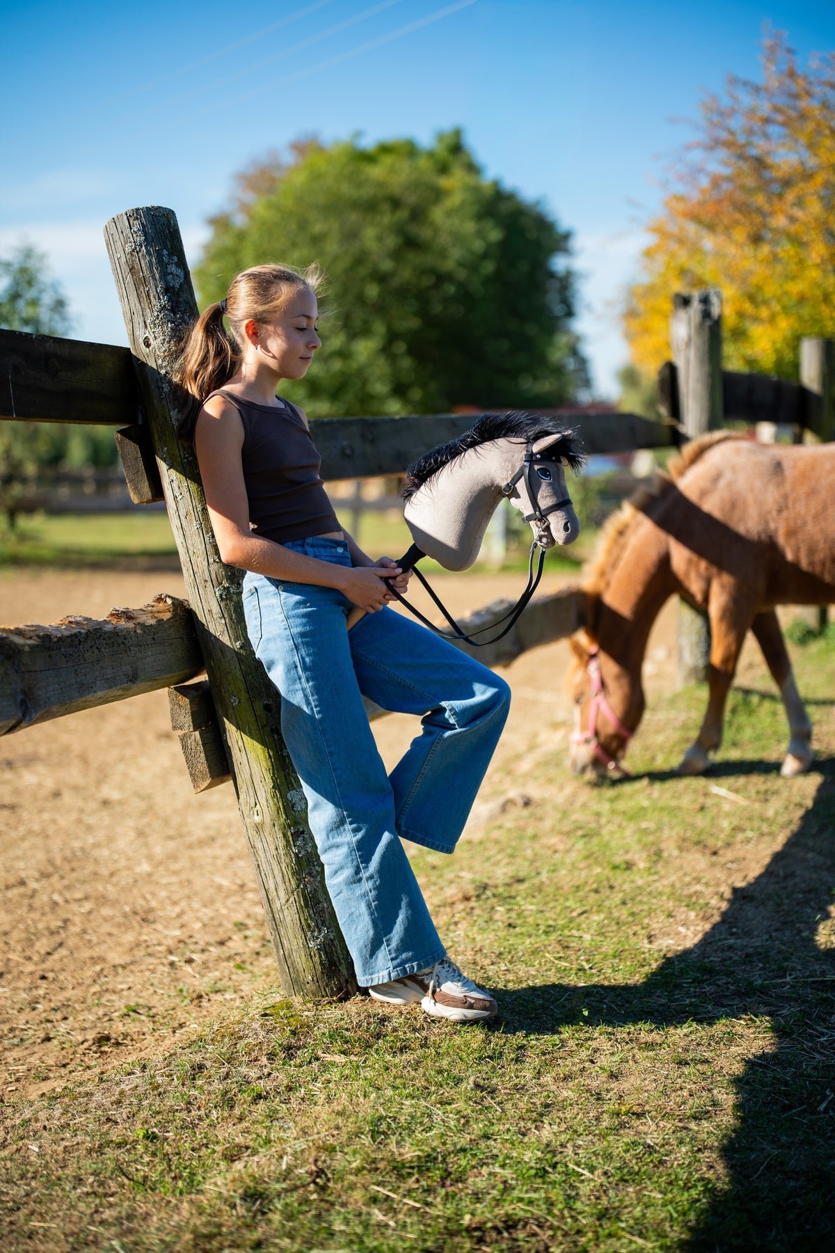 Deklica se v jesenskem soncu naslanja na ograjo izpusta s Hobby Horse konjem Thor s črno uzdo, v ozadju se pase poni lisjak