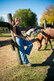 Deklica se v jesenskem soncu naslanja na ograjo izpusta s Hobby Horse konjem Thor s črno uzdo, v ozadju se pase poni lisjak