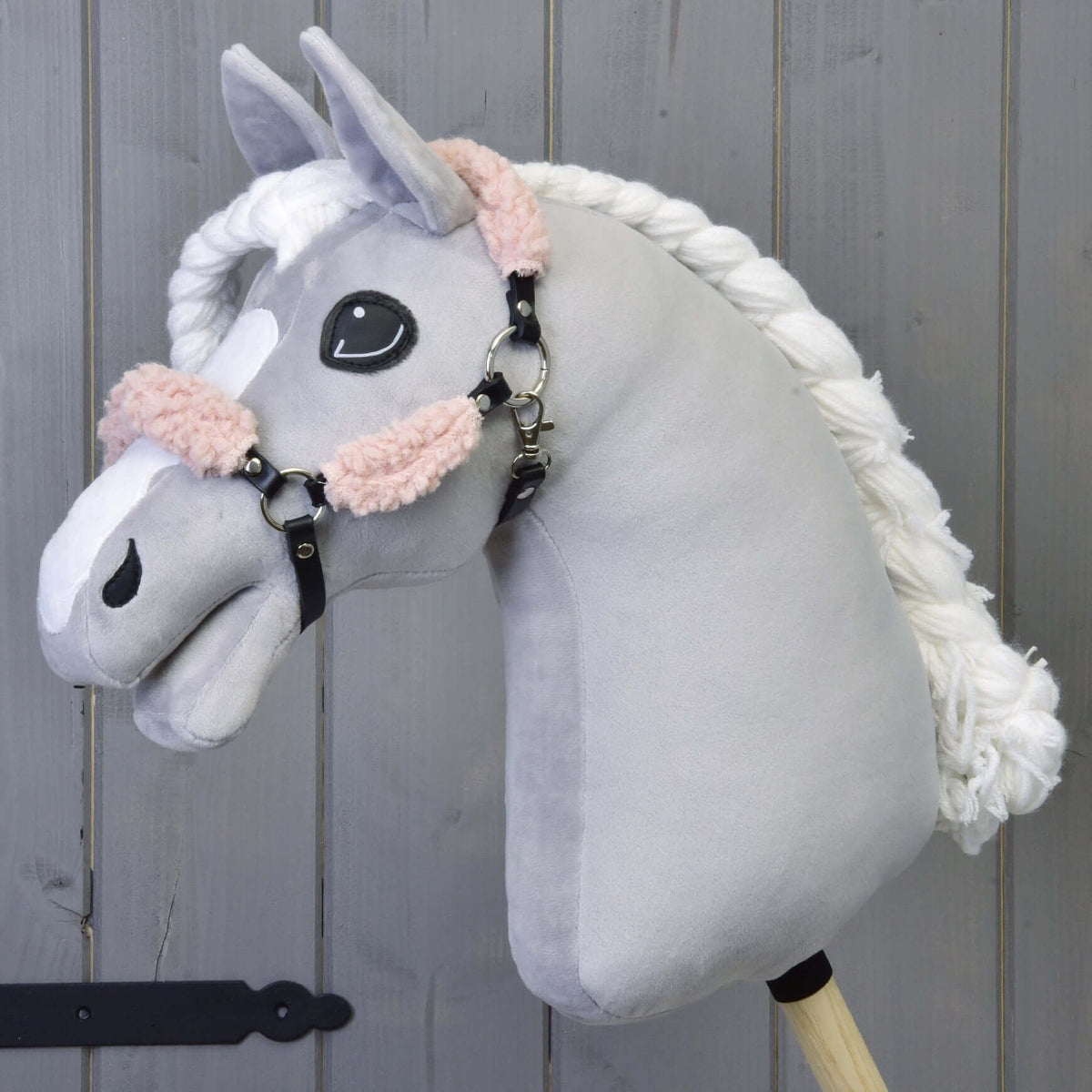 Siv plišast Hobby Horse konj s spleteno belo grivo nosi črno oglavko s staro roza krznom, pred sivo leseno steno