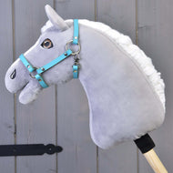 Siv plišast Hobby Horse konj z belo grivo in rjavimi očmi nosi turkizno oglavko iz umetnega usnja, ob sivi leseni steni